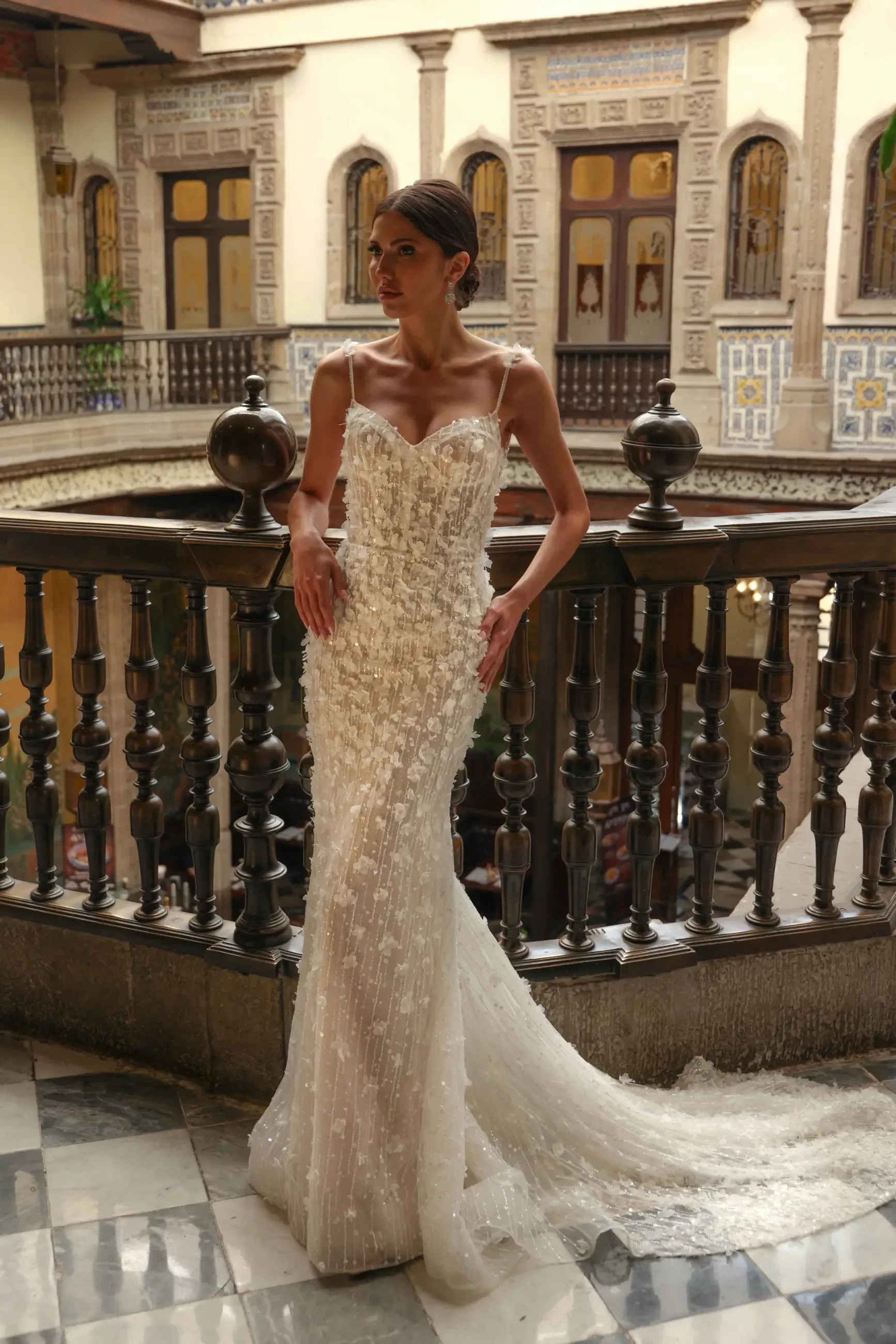 A woman in an elegant, textured white gown stands by an ornate wooden railing in a historic building with arched doorways, conveying elegance and grace.