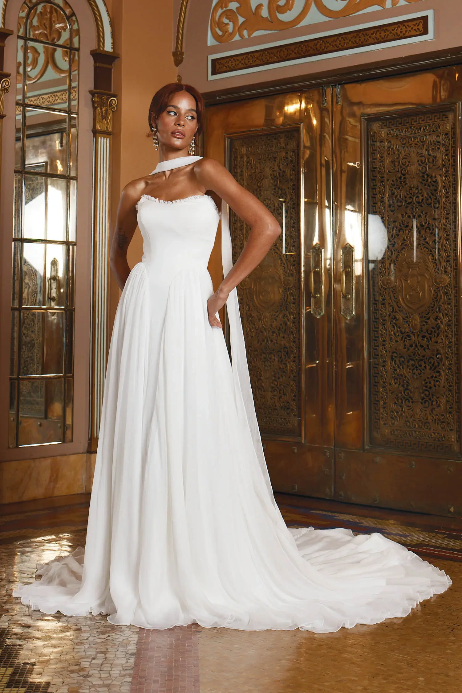 A model wearing a white wedding gown stands in an ornate room with golden doors and decorative mirrors.