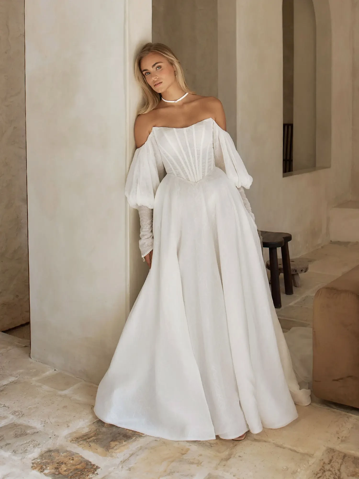 A woman in a white off-the-shoulder dress with puffed sleeves, standing elegantly against a wall in an interior setting.
