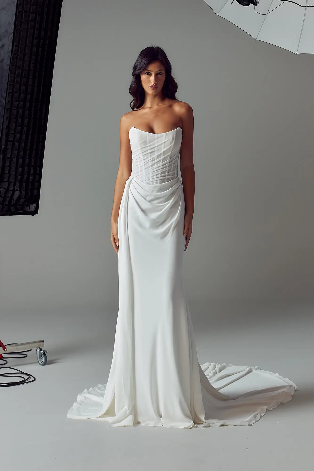 A model wearing a strapless white dress with a fitted bodice and flowing skirt, standing in a photo studio.