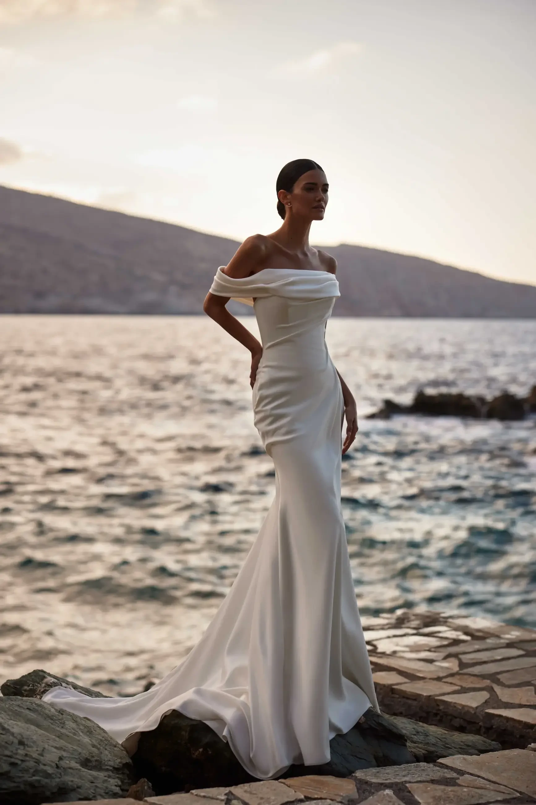 A woman in an off-shoulder, elegant white gown stands by a rocky shoreline at sunset, conveying elegance and tranquility against a scenic ocean backdrop.