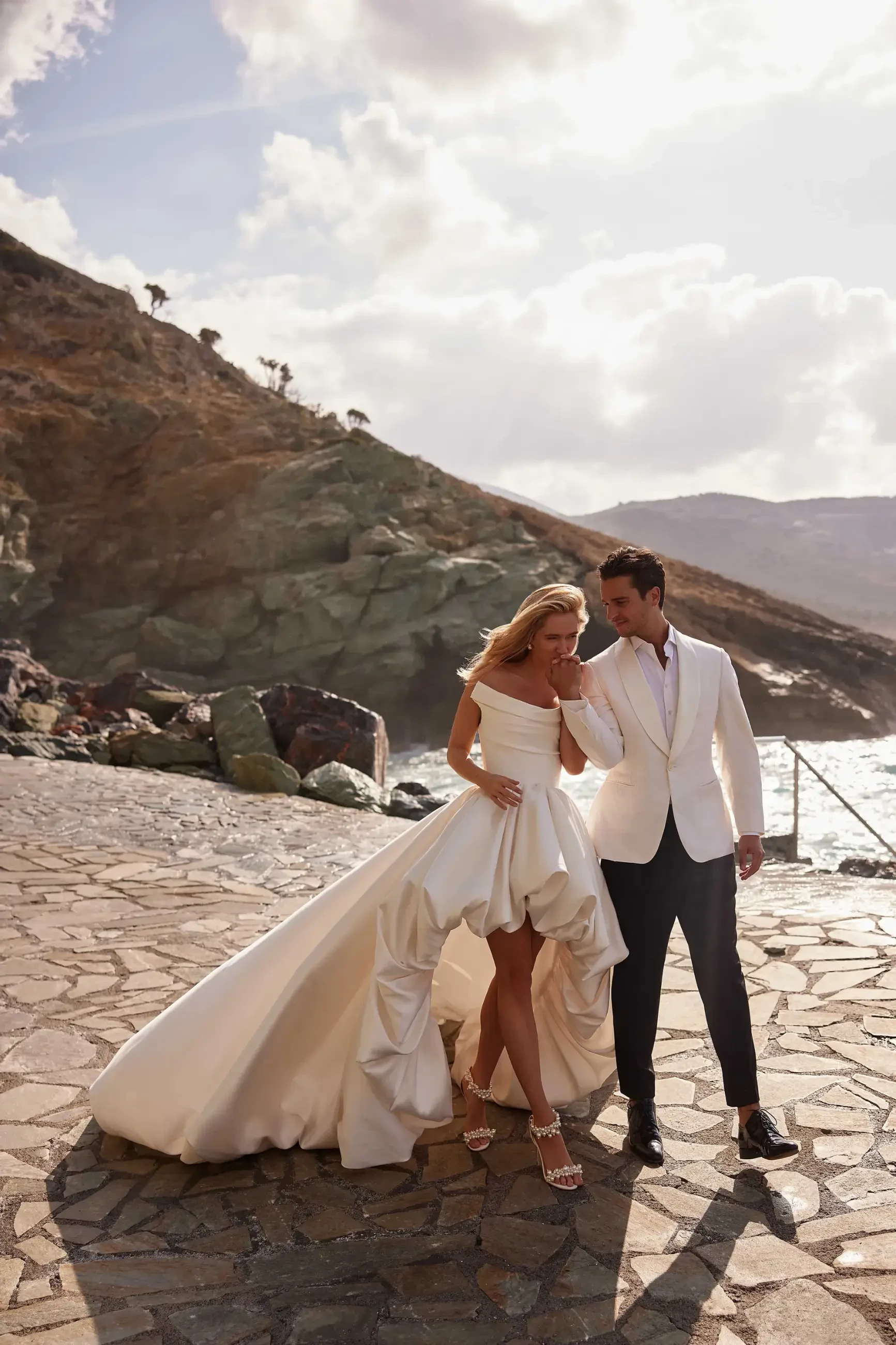 A couple walks along a stone path by the sea, dressed in elegant wedding attire. The bride's gown flows, and the groom wears a white jacket. The scene is sunny with a rocky hillside in the background, creating a romantic and serene atmosphere.