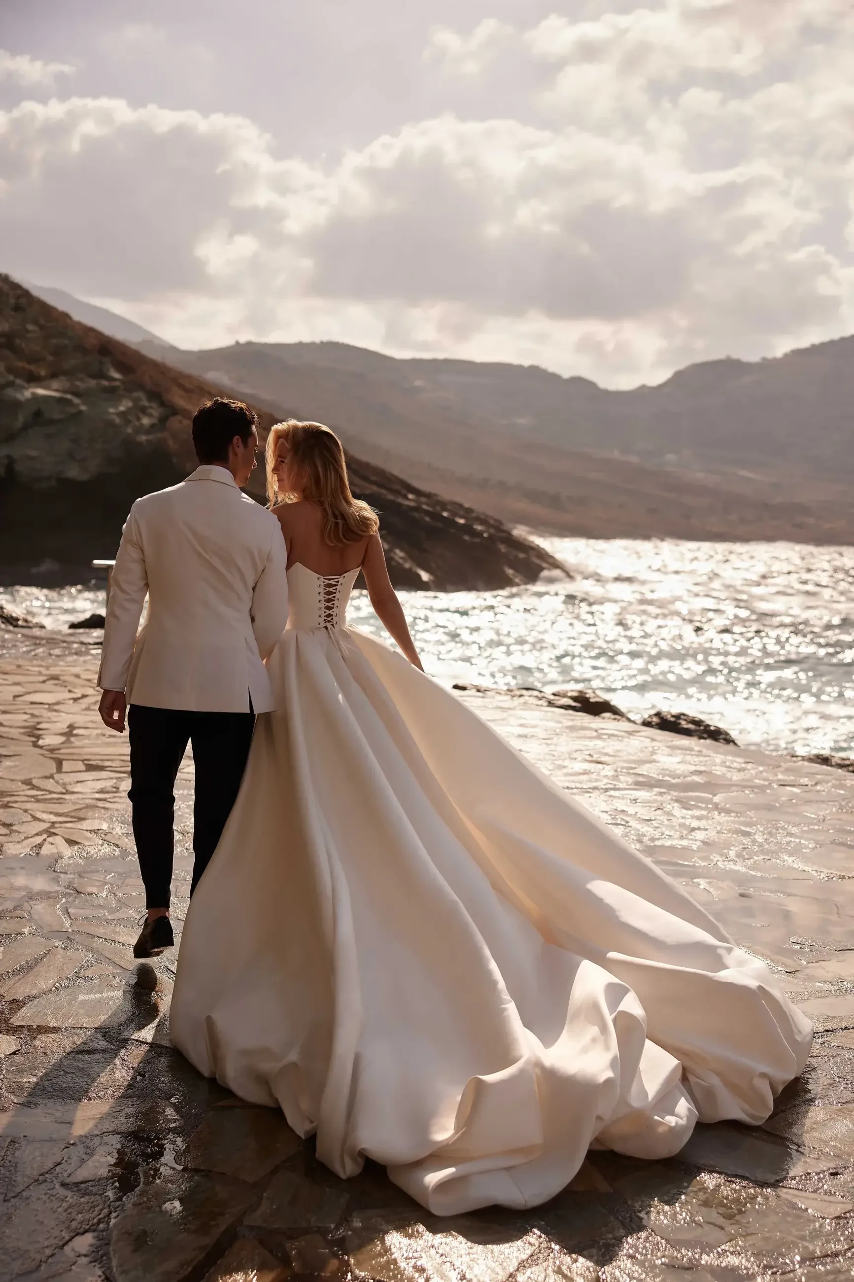 A couple walking hand in hand along a rocky shoreline, with the woman wearing a flowing white wedding dress and the man in a formal white suit, against a backdrop of mountains and water under a cloudy sky.