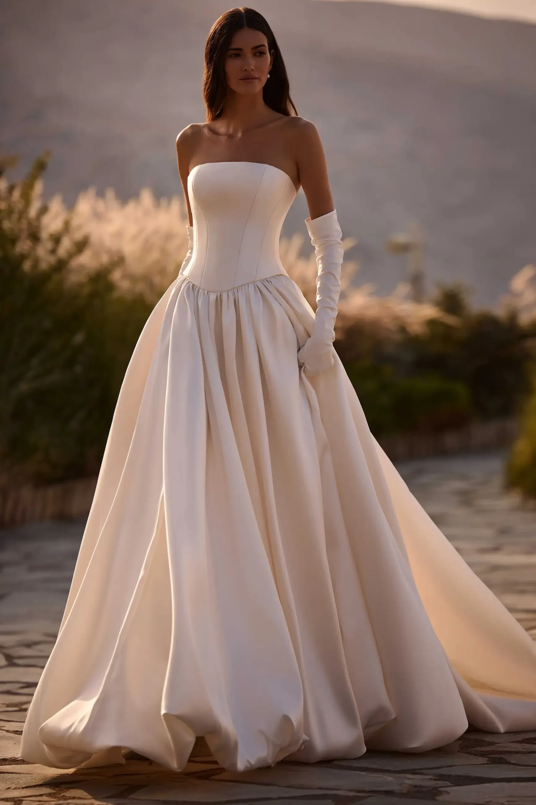 A model wearing a strapless white gown with a full skirt and long gloves, standing outdoors with a blurred background of foliage and soft sunlight.