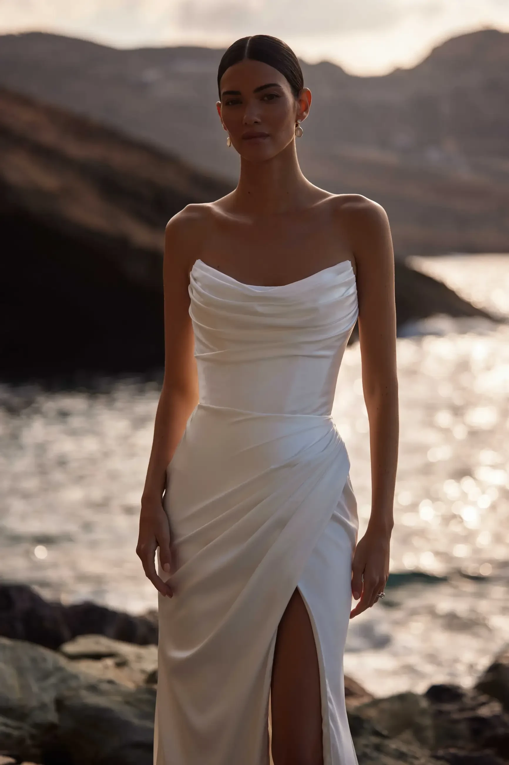 A woman wearing a white dress standing near the water with a rocky landscape in the background.