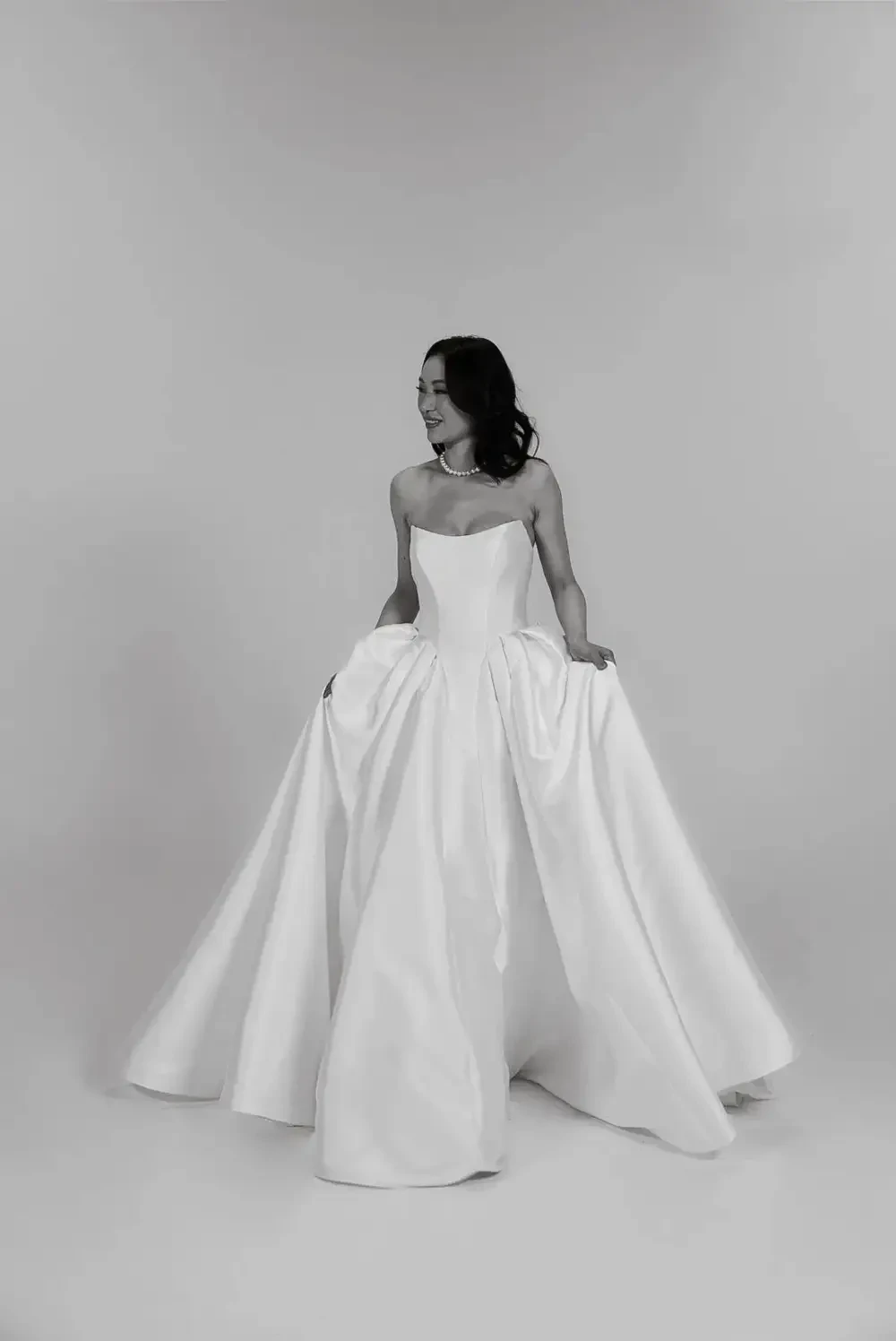 A woman in a strapless white gown stands gracefully, with her hands holding the flowing fabric of the dress. The image is in black and white, highlighting the elegance of the gown.