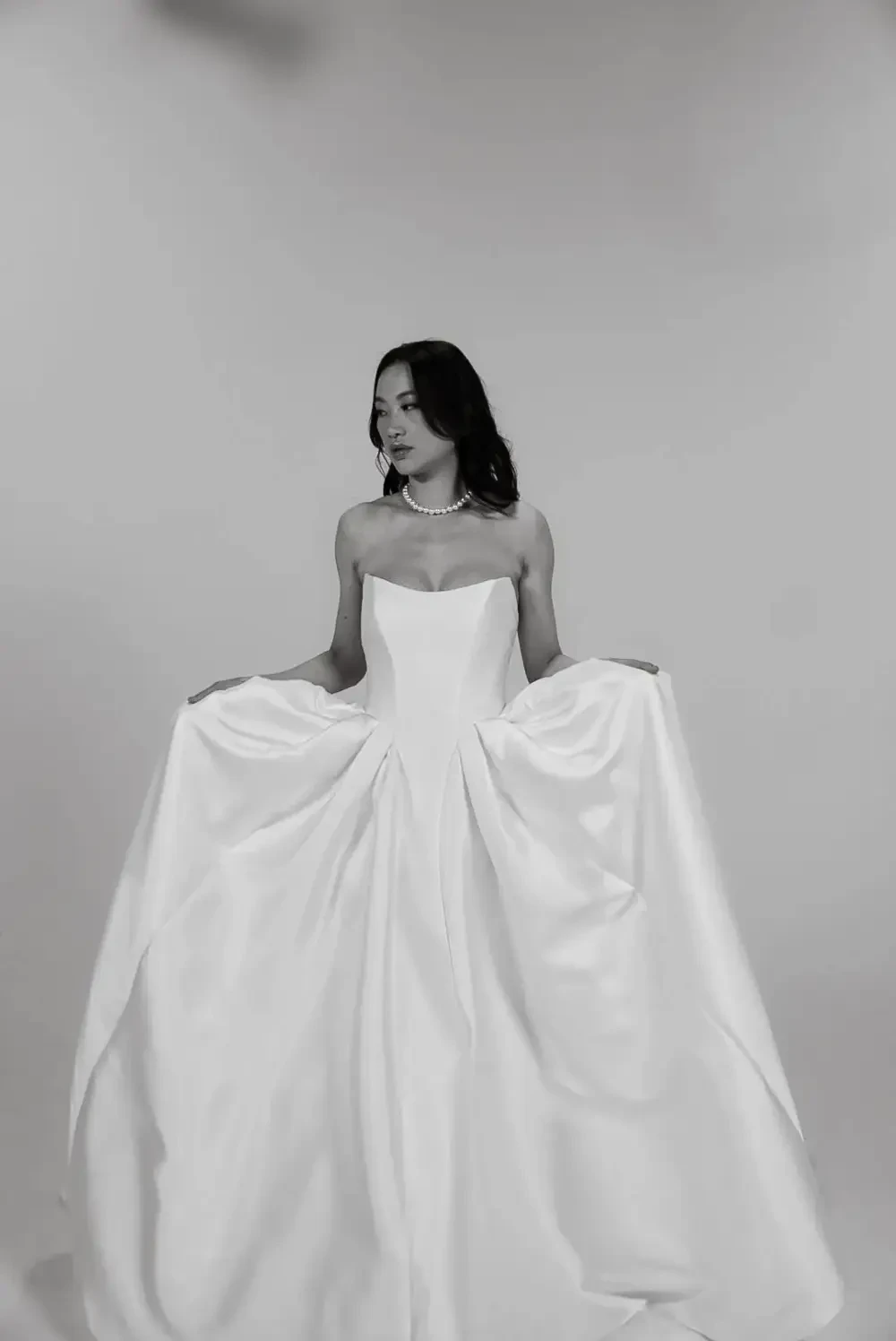 A woman in a white strapless gown, holding the skirt of the dress, with a simple pearl necklace.