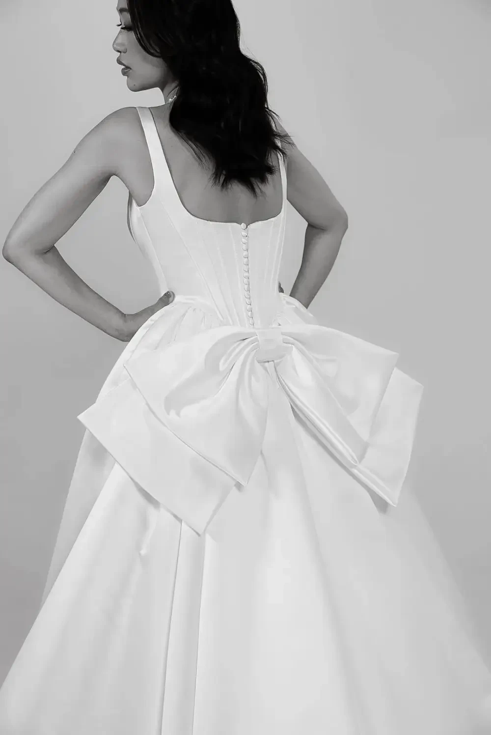 image shows a model wearing a sleeveless bridal gown with a bow detail on the back