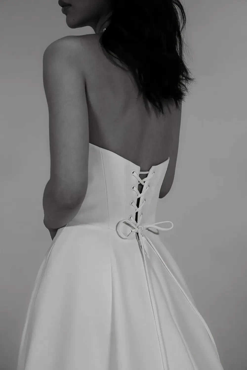 image shows a model wearing a sleeveless bridal gown with a bow detail on the back