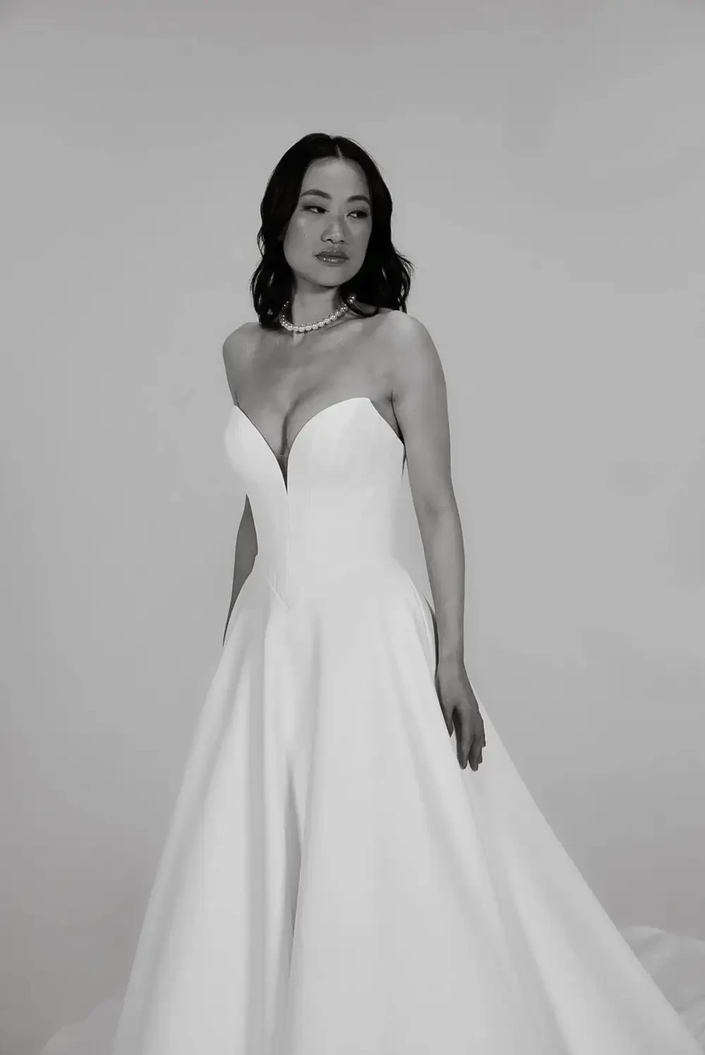 A woman wearing a strapless white wedding dress, standing against a plain background. She has wavy dark hair and is adorned with a necklace, striking a confident pose.