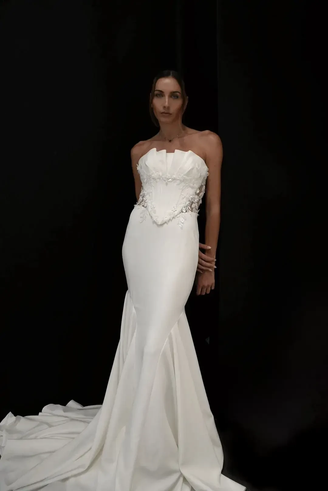 A woman stands confidently in an elegant, strapless white gown with intricate floral detailing on the corset. The long, flowing skirt creates a timeless, graceful silhouette against a dark background.