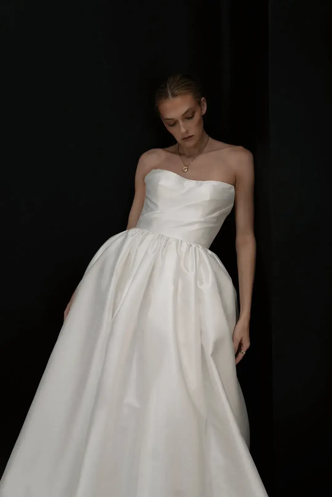 A woman in a strapless white wedding gown stands against a dark background. The elegant satin dress is full-skirted, conveying a serene, contemplative mood.