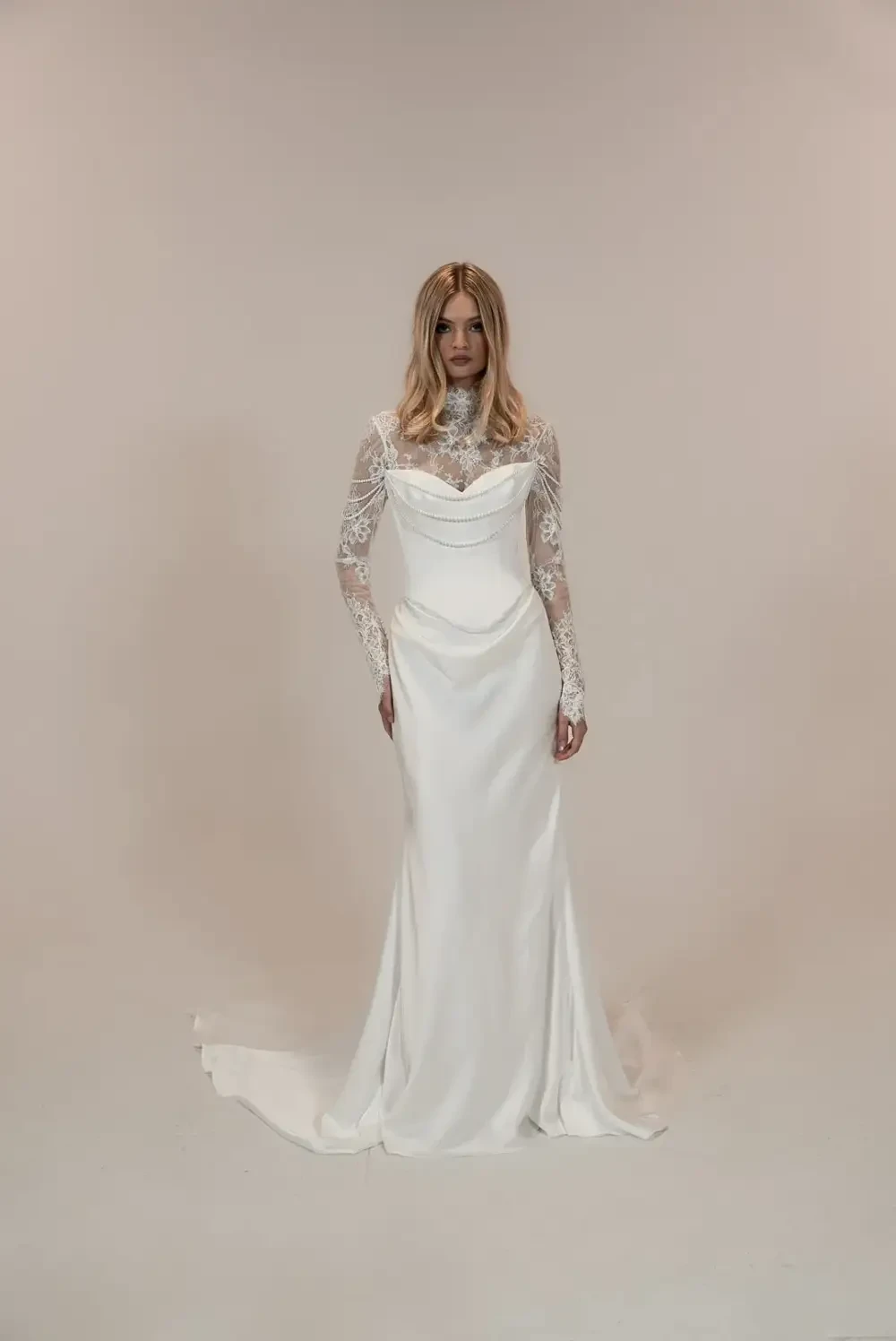 A model wearing a white wedding dress with long lace sleeves, standing against a plain background.