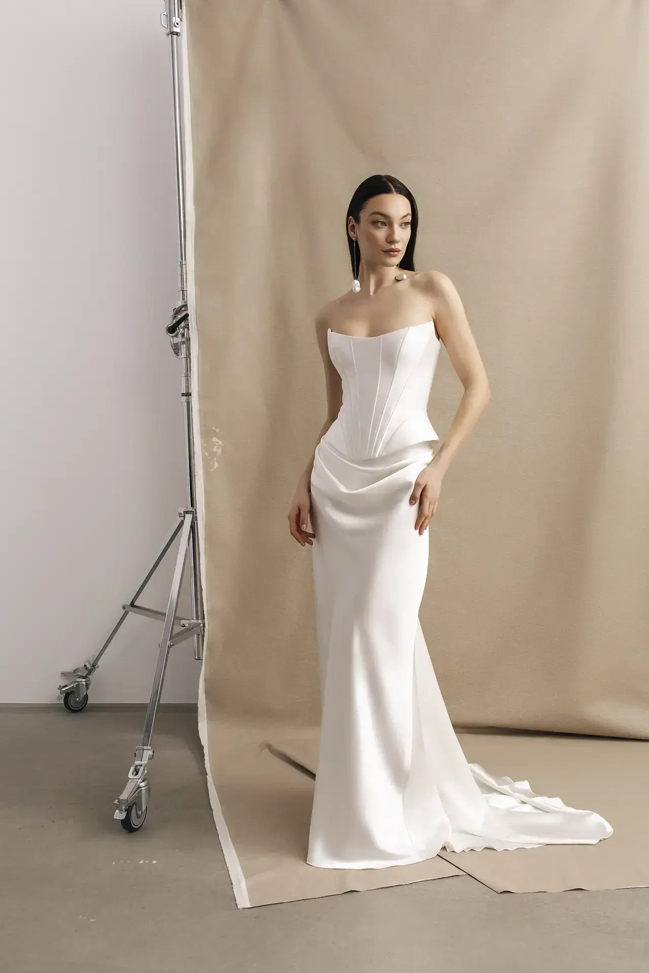 A model stands wearing a sleek, off-white strapless gown with a fitted bodice and elegant draping. The background features a neutral beige color, and the setting appears to be a studio with equipment visible.