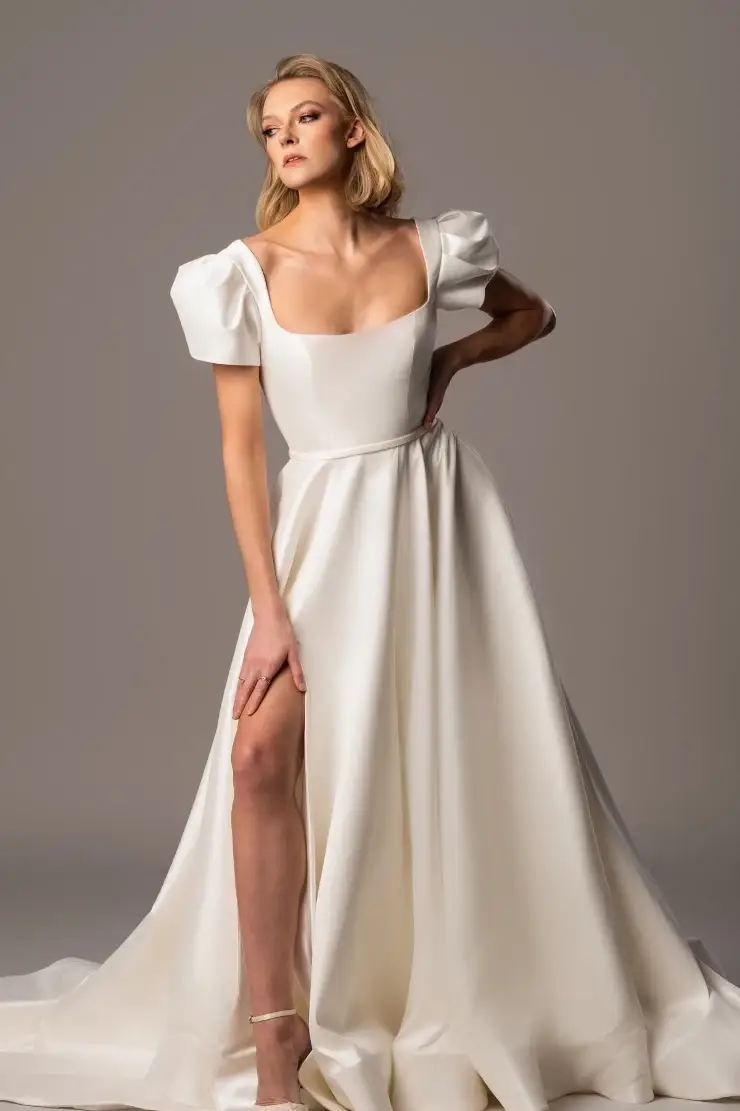 A model poses in a long, elegant white dress with puffed sleeves and a thigh-high slit. The dress has a square neckline and flows gracefully to the floor.