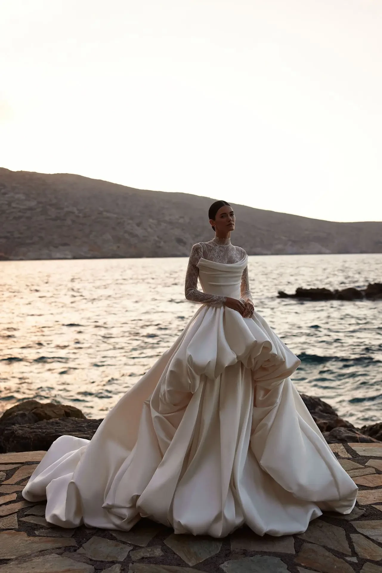 A woman in an elegant, flowing white dress stands by the water's edge at sunset, with mountains in the background.
